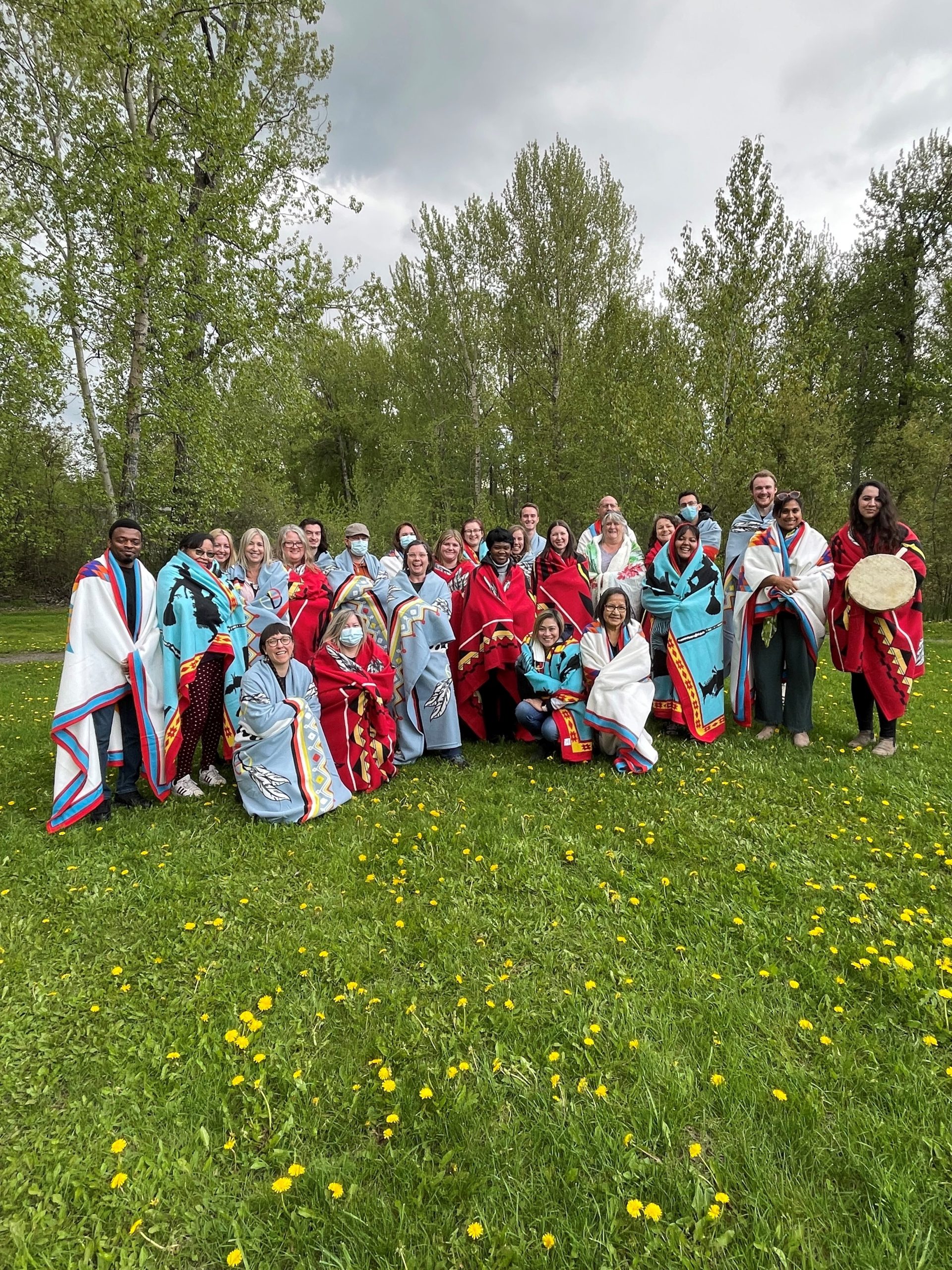 A group of people stand in a flower-dotted field wearing colourful blankets and holding drums.