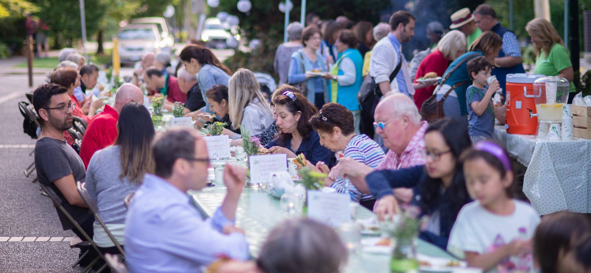 People of all ages sit at a long table outdoors eating food.