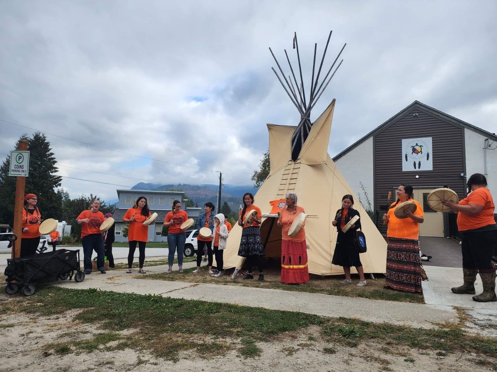 A group of 12 children and adults in orange shirts stands in a line in front of a tipi, holding drums and singing