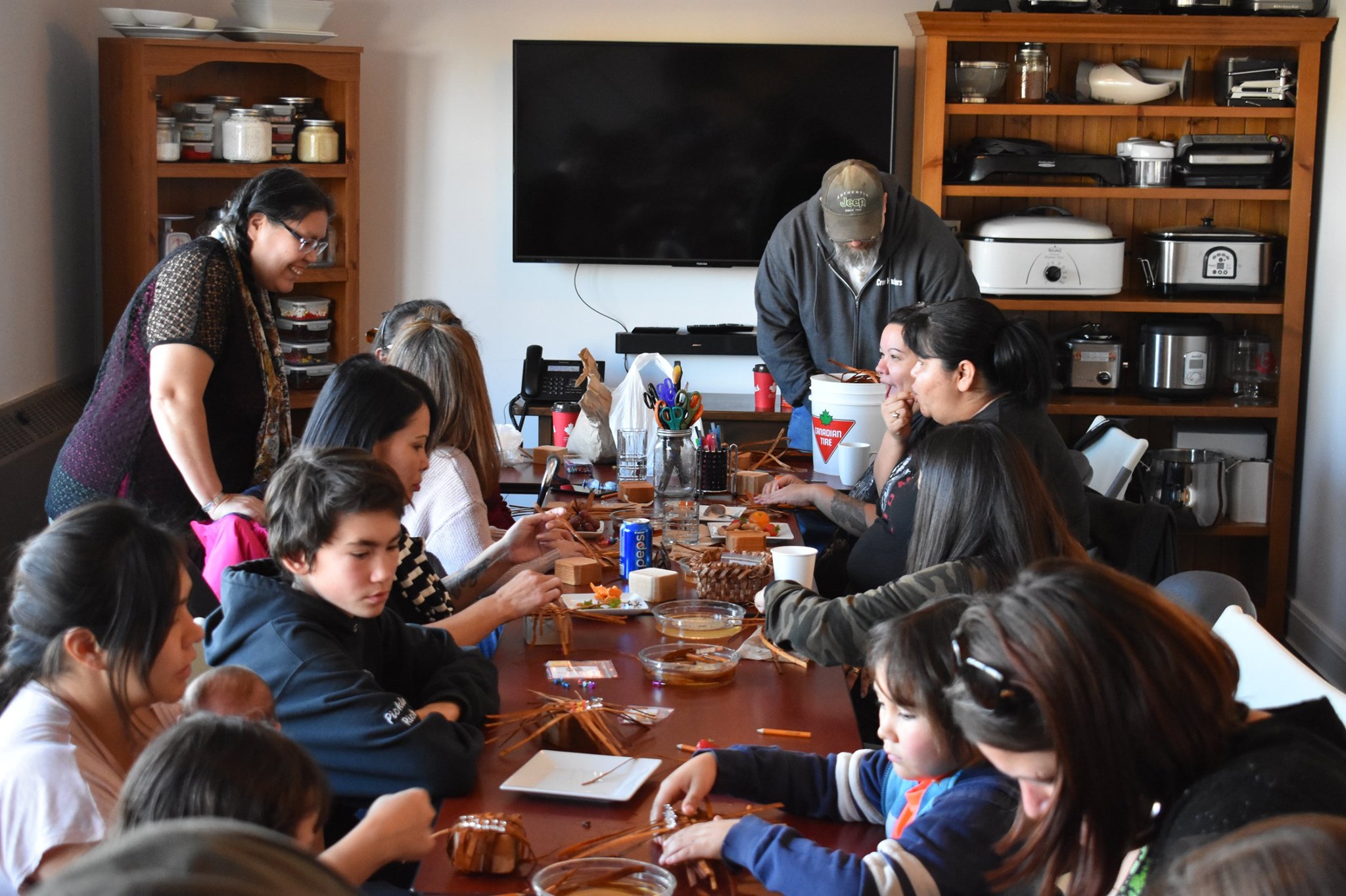 A group of families sits at a table doing cedar weaving.