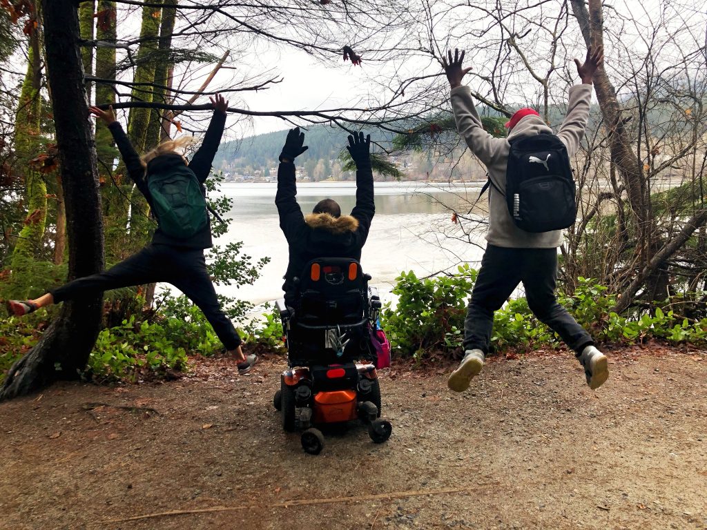 Three people with their backs turned to the camera raise their arms in joy. They are facing a mountain and water scene outdoors.